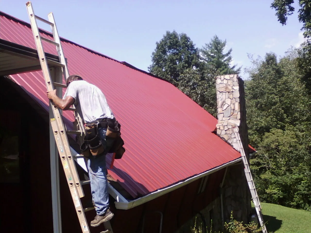 Roofer on a ladder beside a red metal roof and stone chimney — Soffit & Fascia Repair by Roofing Brothers in Bellwood